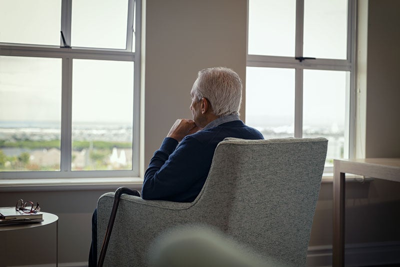 Elderly man from behind, sitting in armchair, gazing thoughtfully out of a large window at a town beyond.