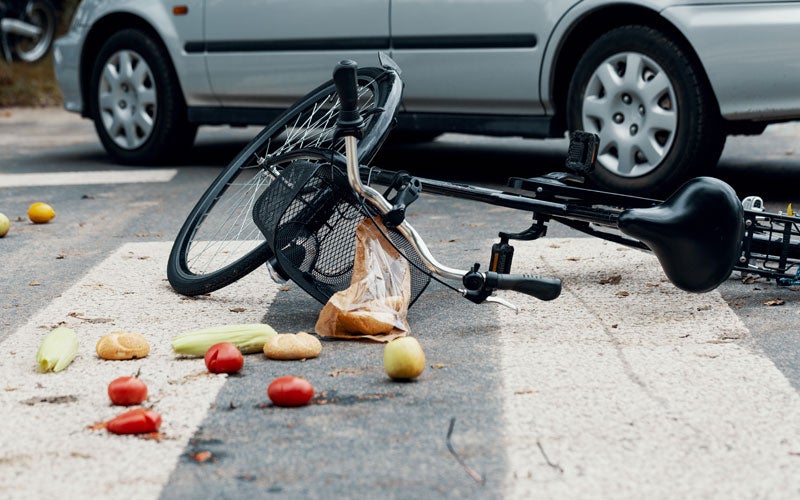 A black bicycle with groceries including tomatoes, limes, and apples scattered on a crosswalk near a silver car.