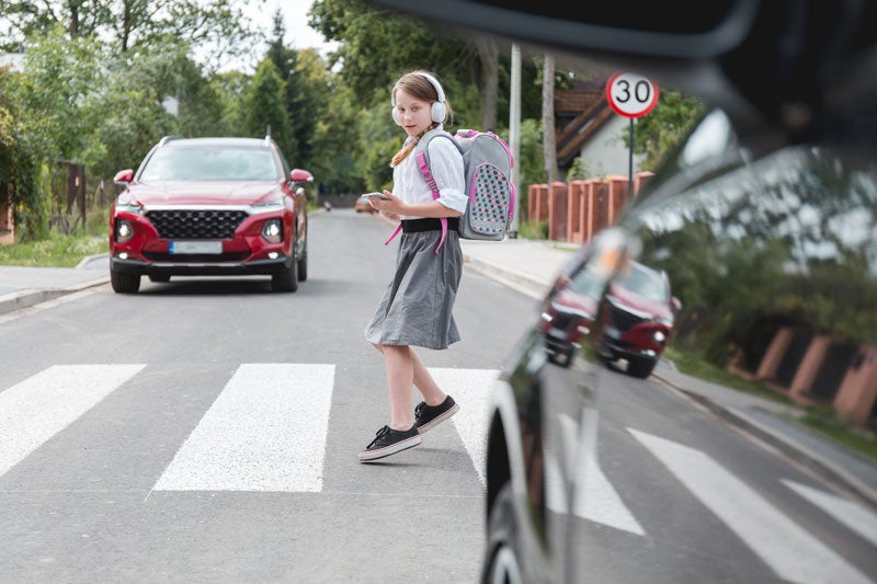 A girl with headphones and backpack looking at her phone while crossing a street in front of a red car.