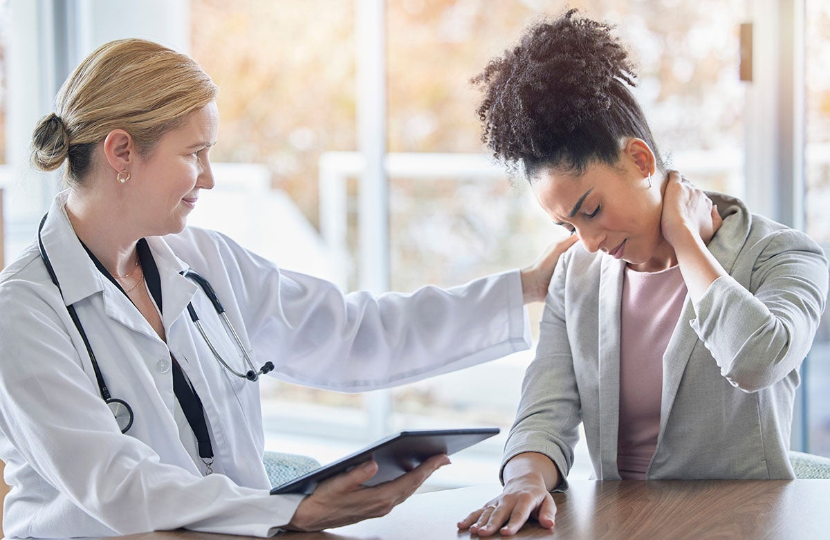 A female doctor in a white coat holds a tablet and comforts a female patient with a painful neck.