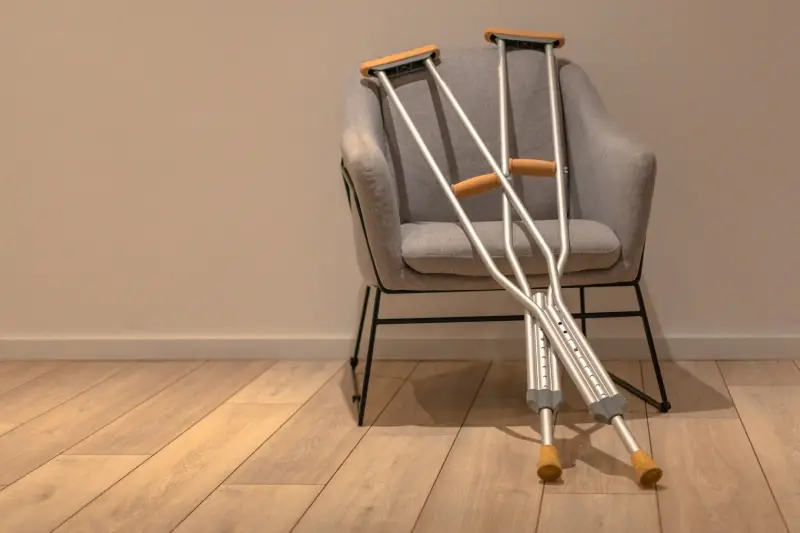 Crutches leaning against a gray armchair on a wooden floor, with a light-colored wall in the background.