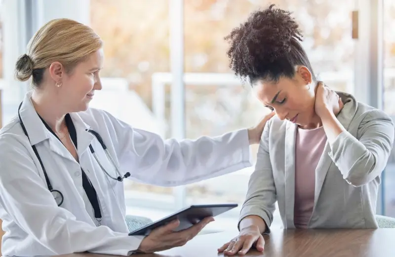 A caring doctor touches the shoulder of a patient who is clutching her neck in pain.