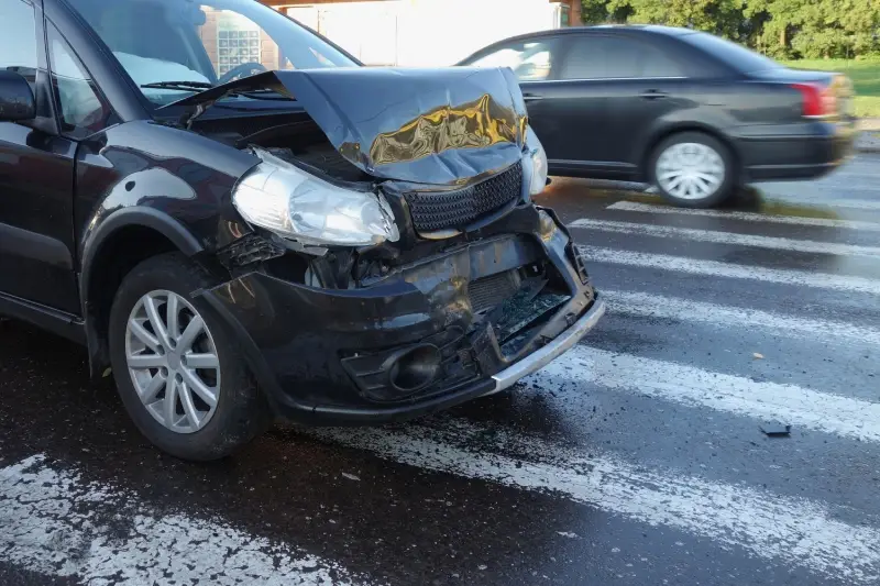Front of a dark car with a crumpled hood and damaged bumper parked on a wet crosswalk.