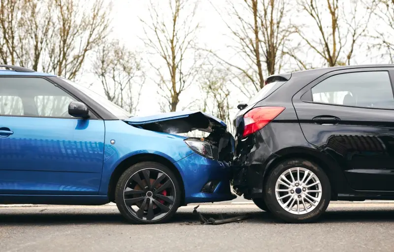 A blue car crashed into the back of a black car, both severely damaged on the road, with trees in the background.