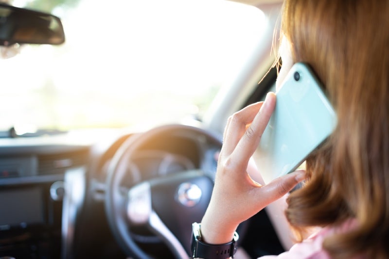 Person with brown hair talks on a light blue smartphone while driving, with a glimpse of the steering wheel.