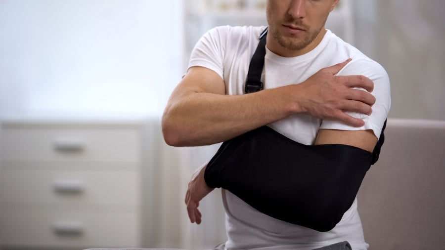 A man in a white t-shirt wearing a black arm sling supports his injured shoulder with his other hand.