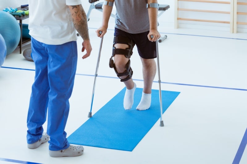 A physical therapist guides a person with a knee brace and crutches walking on a blue mat in a gym.