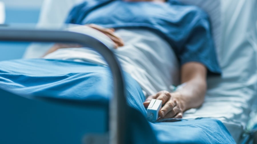 A patient in a blue hospital gown lies in a hospital bed, an IV in their arm and a pulse oximeter on their finger.