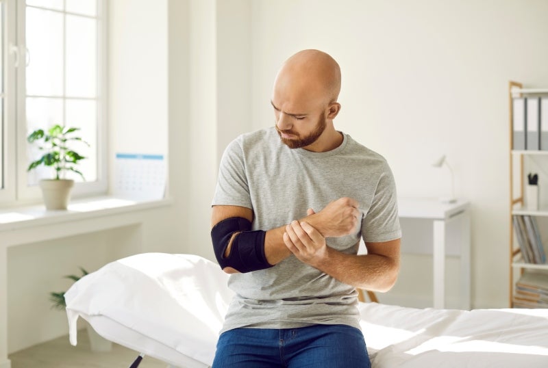 A man with an elbow brace holds his wrist, looking down in discomfort, sitting on an examination table.