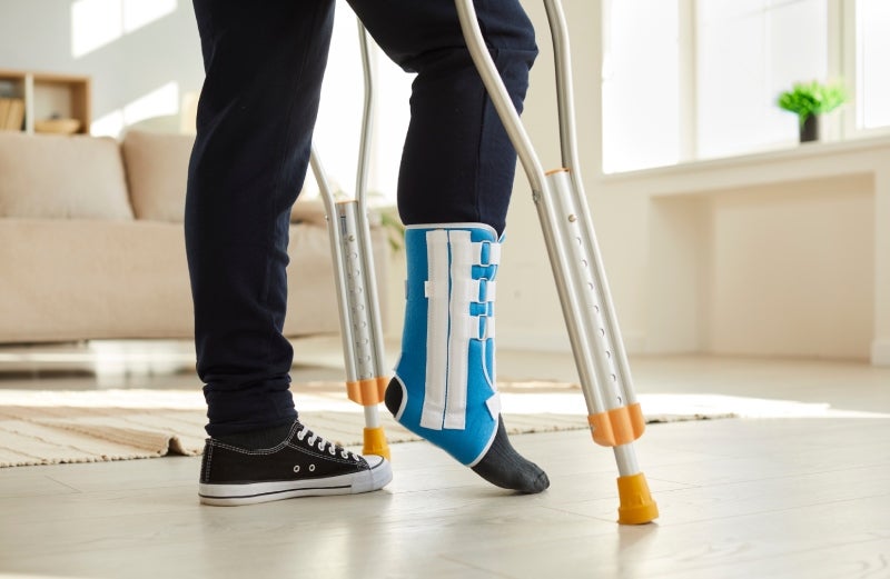 A person with a blue foot brace and dark socks on one foot, wearing a black sneaker on the other, walks with crutches.