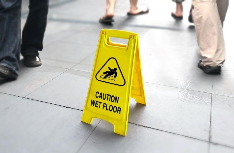A yellow CAUTION WET FLOOR sign with a slipping person symbol on a tiled floor with blurred people walking.