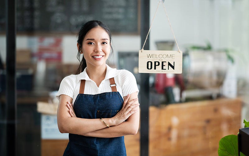 A smiling young woman in a denim apron stands confidently in front of a glass door with an 'OPEN' sign.