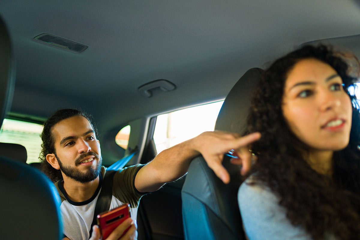 A man in the back seat of a car holds a red phone and gestures toward a woman in the front seat.