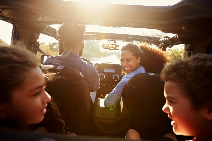 Family of four, including two adults and two children, enjoying a sunny road trip in a car.