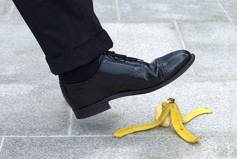 A foot in a black dress shoe is about to step on a banana peel on a grey tiled surface.