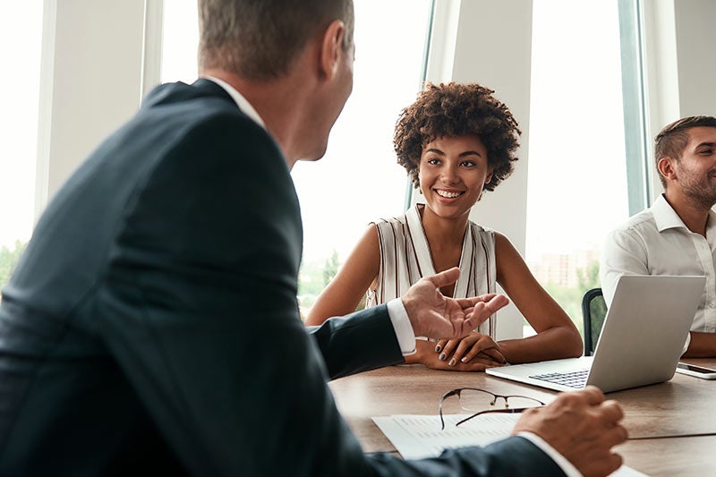 A smiling Black woman in a striped top looks at a gesturing man in a suit across a conference table.