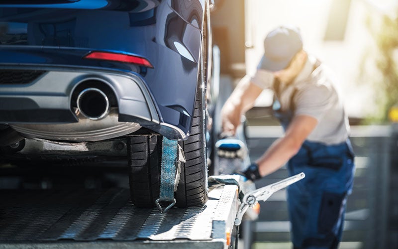 Rear view of a blue car being loaded onto a tow truck, with a blurred driver in the background.