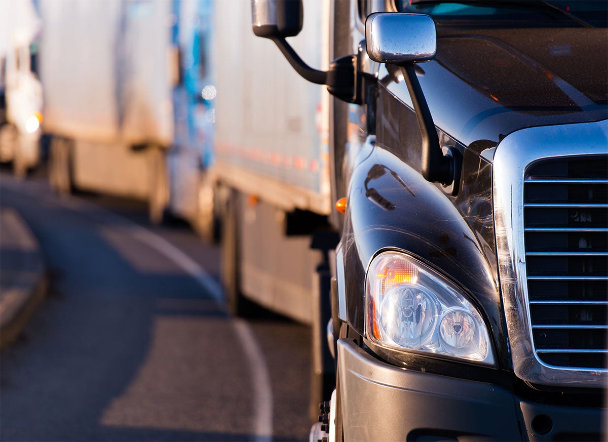 Front of a black semi-truck in the foreground, with a line of other semi-trucks in the blurred background.