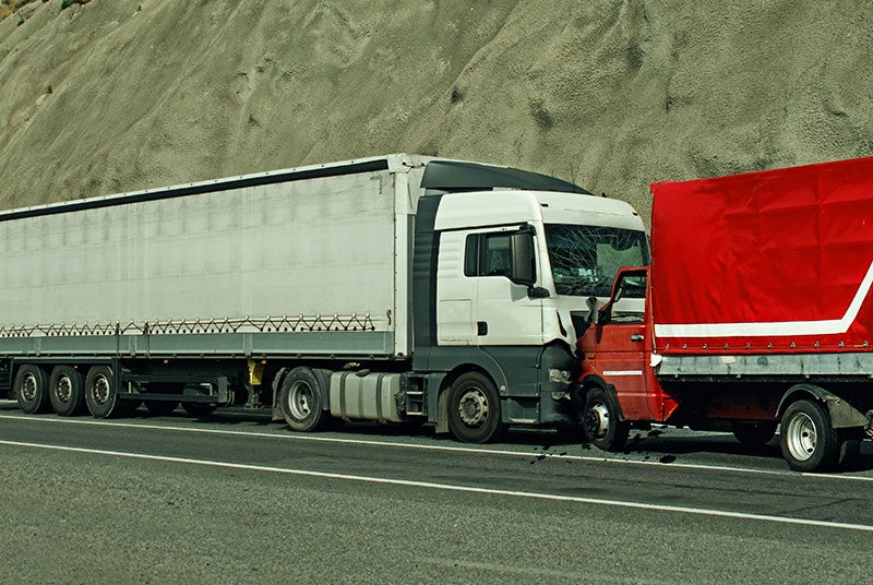 A white semi-truck with a damaged windshield after colliding with a smaller red truck with a matching red trailer.