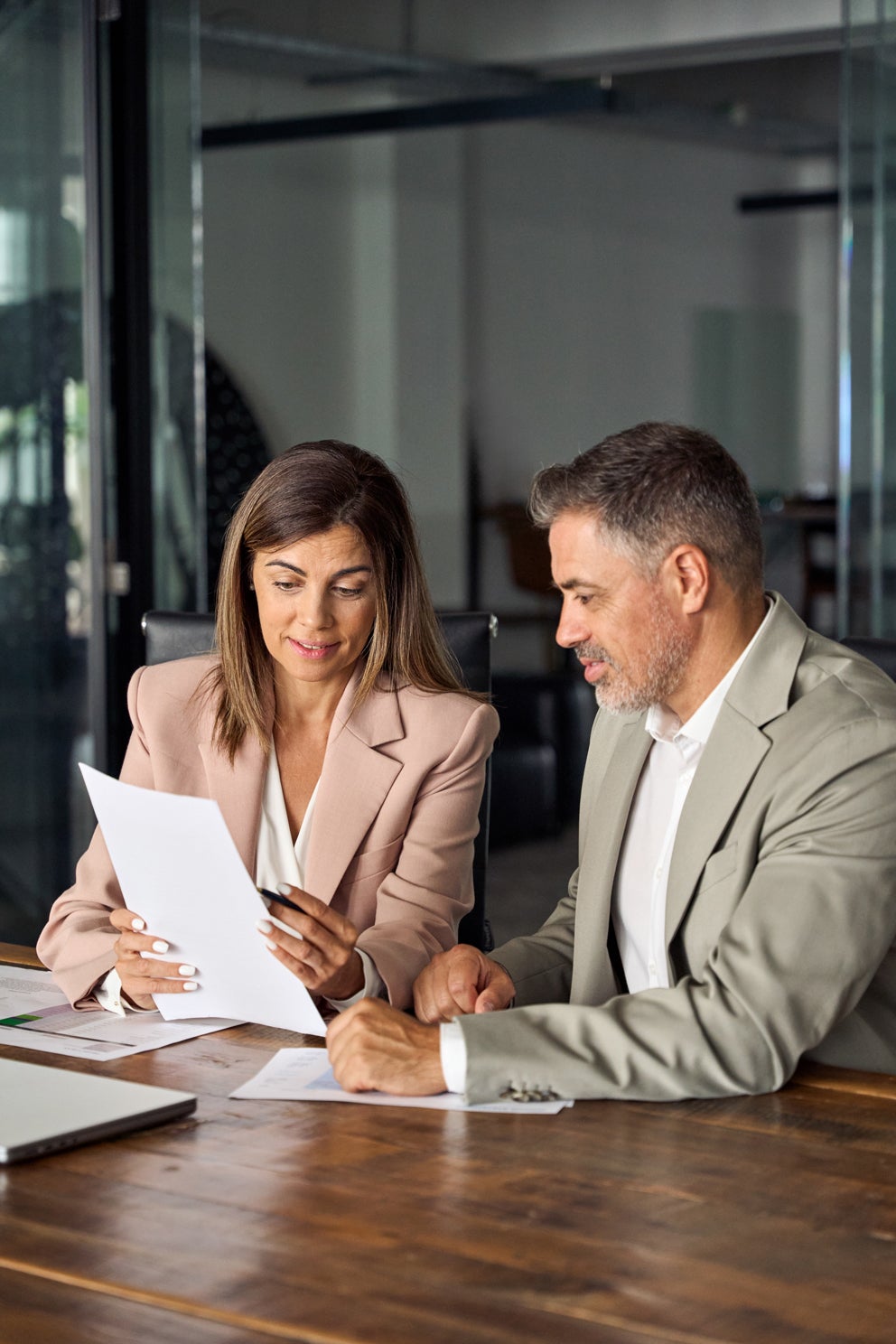 Two elegantly dressed business people, a woman and a man, review documents together at a conference table.