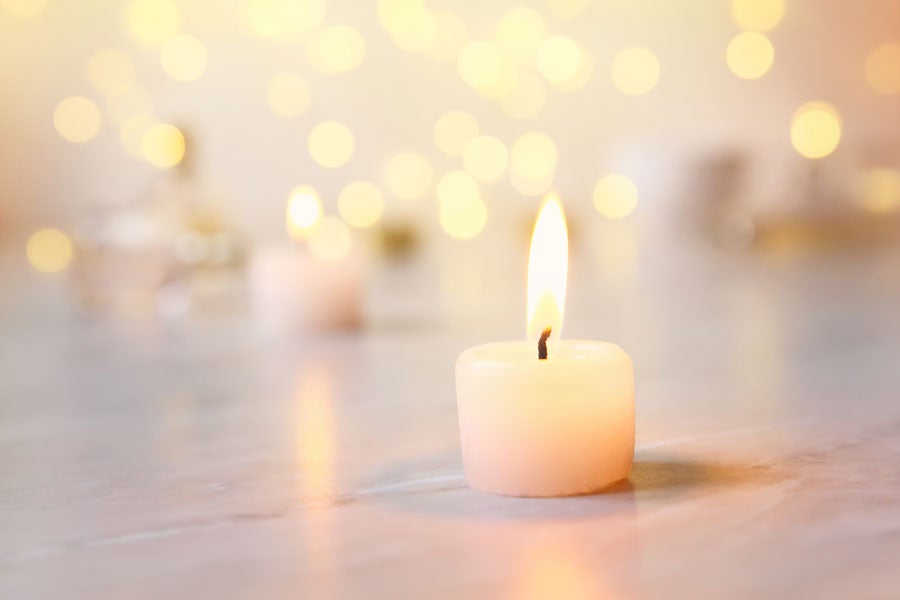 A single lit white tea light candle on a light surface with blurred golden bokeh lights in the background. 