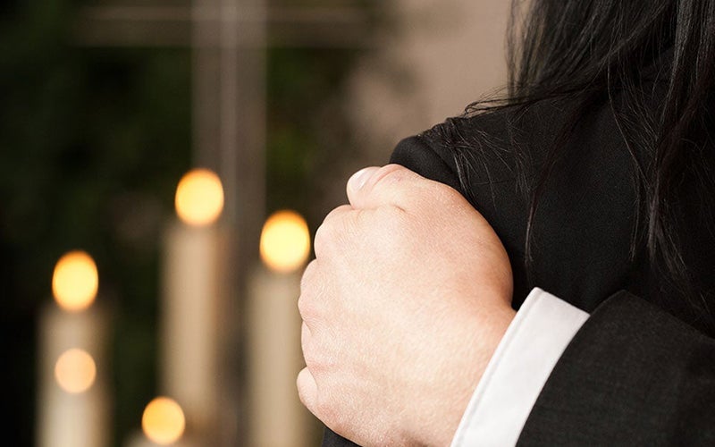 A hand in a white cuff rests on a person's black-clad shoulder, with blurred candle flames in the background.