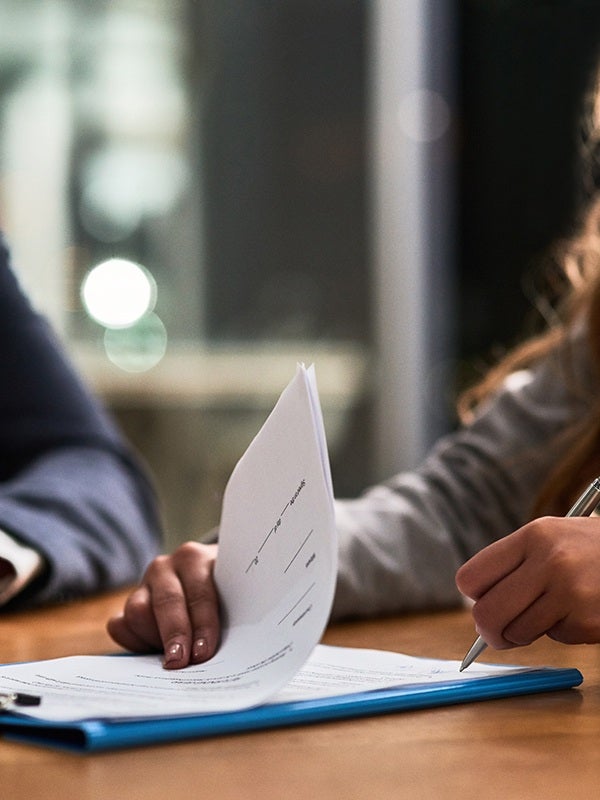 Two people at a table, one flipping through papers and the other signing a document on a blue clipboard.