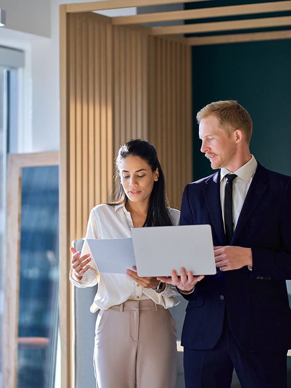 Two business professionals, a woman and a man, review a document and laptop in a modern office.