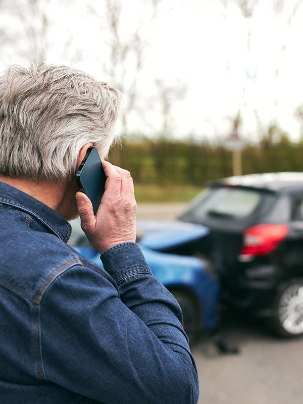 Man with gray hair wearing a denim shirt talking on a phone after a car accident.