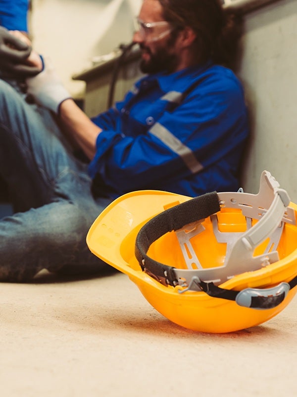 A yellow hard hat on the floor with a worker in a blue uniform and safety glasses sitting against a wall behind it.